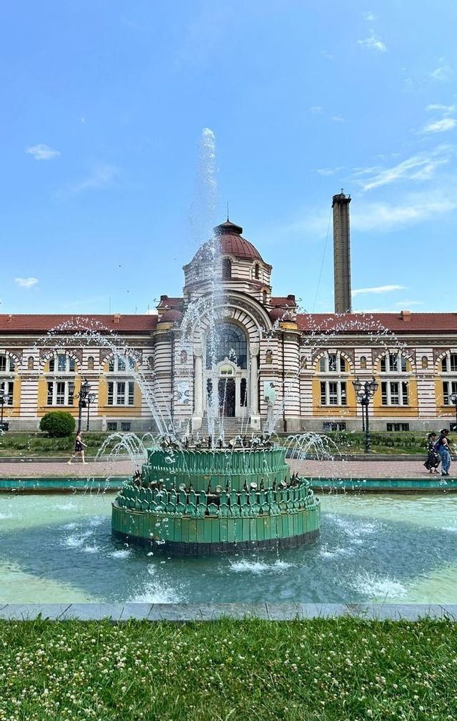 Une fontaine verte à plusieurs niveaux projette de l'eau devant un majestueux bâtiment historique coiffé d'un dôme.