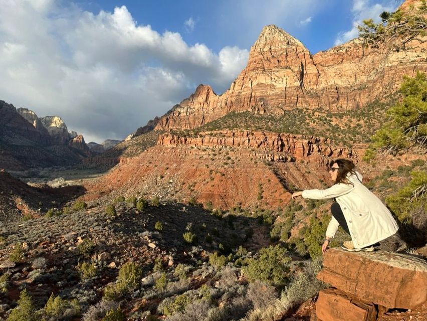 Una mujer con chaqueta blanca se cuclillas en una cornisa de roca con vistas a un vasto cañón de roca roja bajo un cielo parcialmente nublado