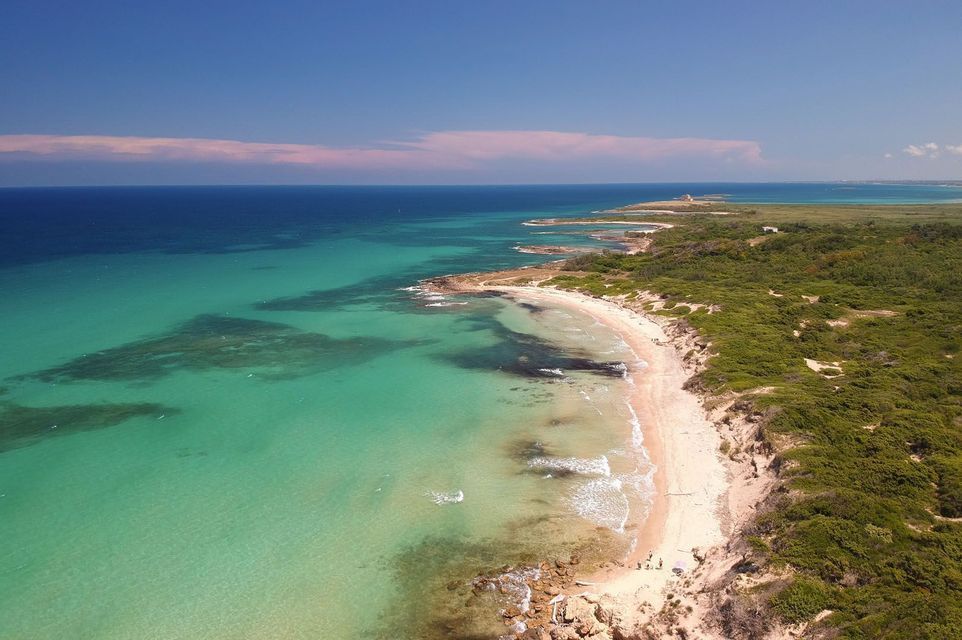 Una vista aérea de una extensa costa curva con una playa de arena, agua turquesa y exuberante vegetación verde bajo un cielo azul claro.