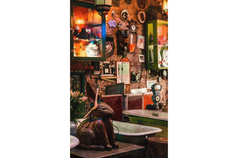 Una grande statua di coniglio si trova su un tavolo in una stanza piena di oggetti d'antiquariato, curiosità e vecchie vasche da bagno, contro un muro di pietra ingombro.