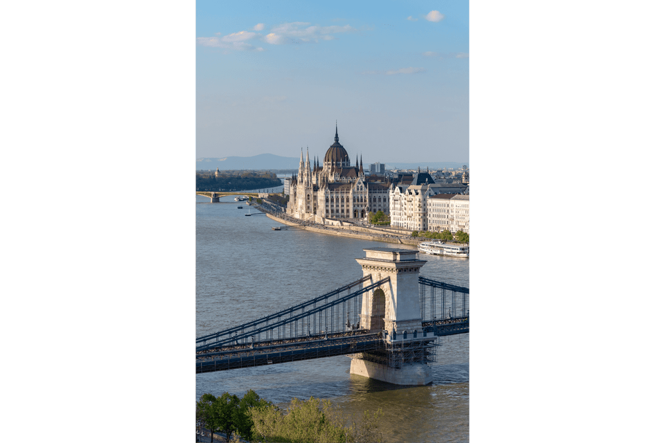 Un ponte a catena storico attraversa un ampio fiume, con un palazzo del parlamento ornato e a cupola sulla riva opposta sotto un cielo blu.