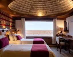 Ein Hotelzimmer mit zwei Einzelbetten und einer gewölbten, strukturierten Decke blickt durch ein großes Fenster auf eine Wüstenlandschaft.