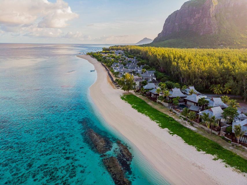 Una vista aerea di un resort su una spiaggia di sabbia bianca, delimitata da acque turchesi e una montagna boscosa sotto un cielo parzialmente nuvoloso.