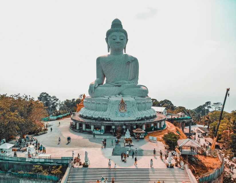 Una vista aerea di una gigantesca statua bianca del Buddha assisa su una collina, con turisti che camminano nella piazza sottostante.