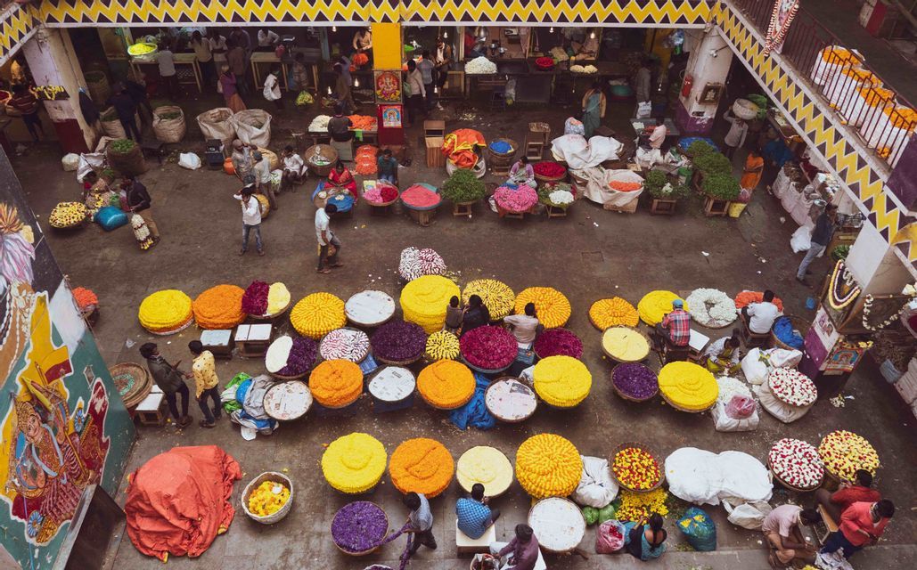 Una vista dall'alto di un vivace mercato coperto ricolmo di grandi cesti rotondi di petali di fiori colorati: gialli, viola e arancioni.
