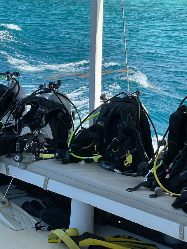 Attrezzatura per immersioni subacquee, incluse bombole e GAV, su una panca di una barca con l'oceano turchese sullo sfondo.