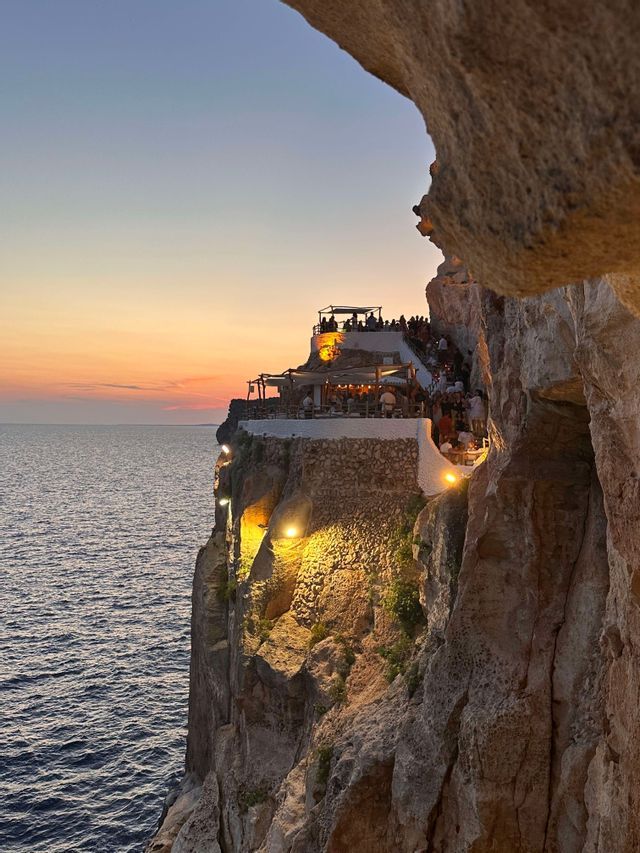 Un ristorante a picco sulla scogliera è illuminato da luci calde che si affacciano sull'oceano al tramonto, visto da una grotta.