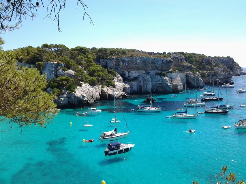 Una vista dall'alto di barche che galleggiano in una cala con acqua turchese, circondata da scogliere rocciose coperte di alberi verdi.