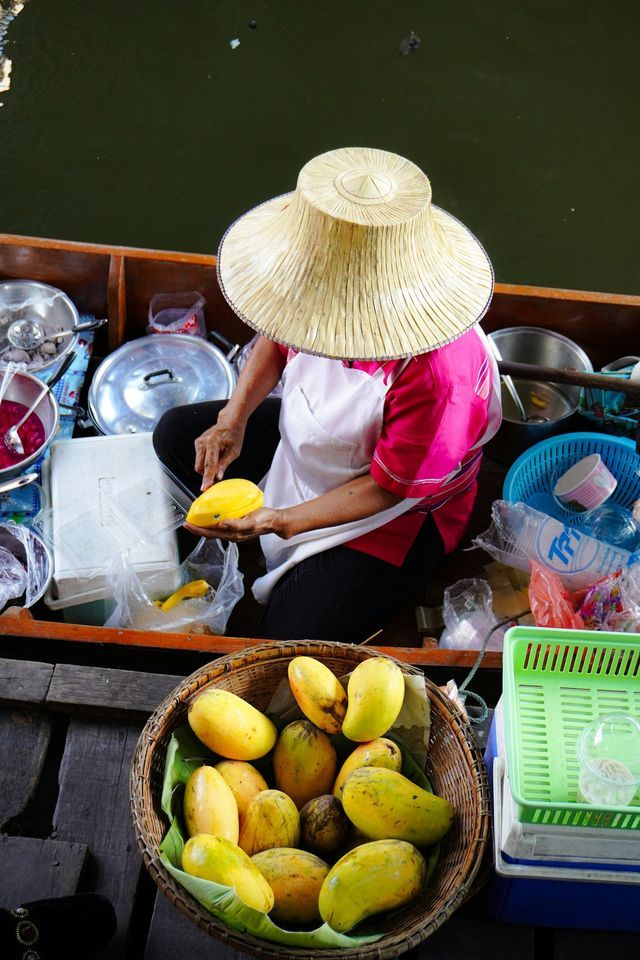 Una vista dall'alto di un venditore con un cappello di paglia che affetta un mango giallo maturo mentre è seduto in una piccola barca.