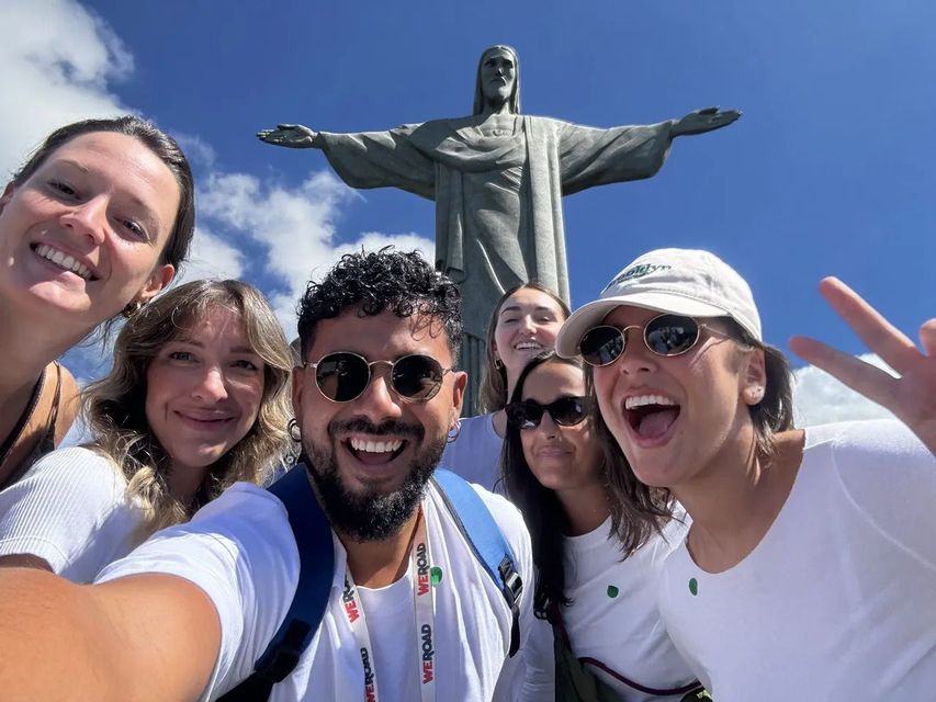 Un gruppo WeRoad in viaggio scatta un selfie sorridente davanti a una grande statua raffigurante una figura con le braccia aperte.