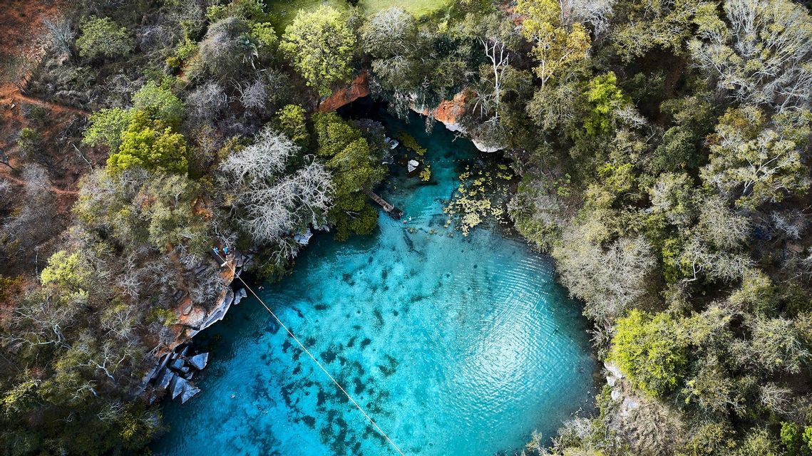Una veduta aerea di un cenote turchese in una foresta, con una zipline che lo attraversa e persone che nuotano nell'acqua cristallina.
