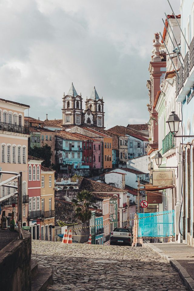 Una vista lungo una ripida strada acciottolata verso colorati edifici storici e una chiesa su una collina sotto un cielo nuvoloso.