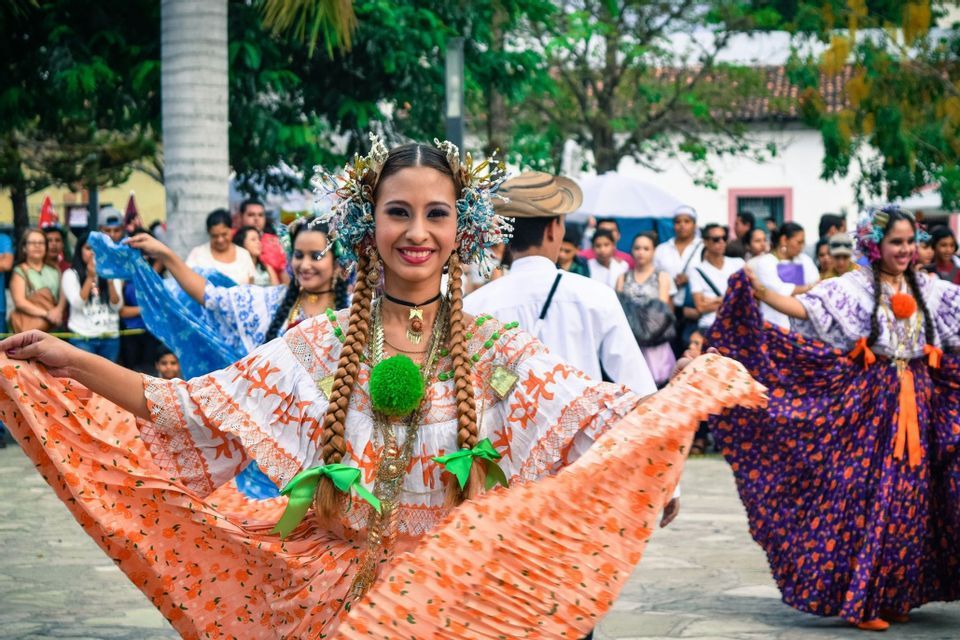 Eine Frau in einem farbenfrohen traditionellen Kleid und Kopfschmuck lächelt, während sie bei einer Freiluftparade tanzt.