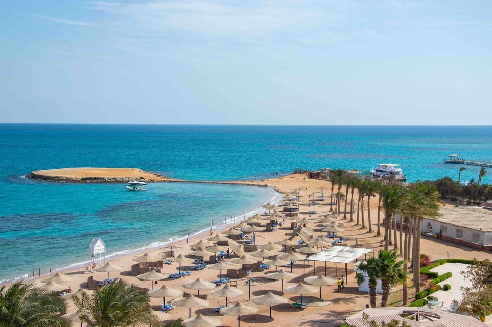 Vista dall'alto: spiaggia sabbiosa, palme, ombrelloni di paglia e mare turchese con isolotto.