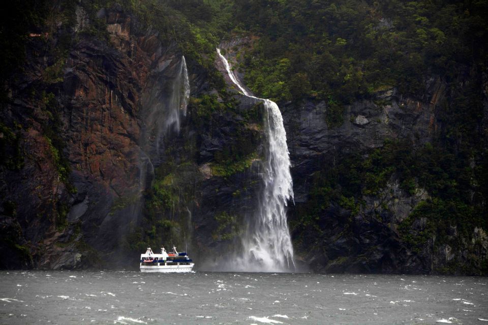 Un'imbarcazione turistica naviga in acque agitate ai piedi di due potenti cascate che si riversano da una scogliera rocciosa e lussureggiante.