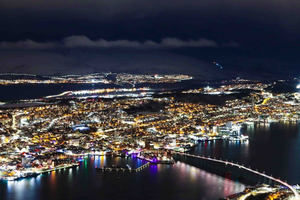 Veduta aerea di una città costiera innevata illuminata di notte, con un ponte illuminato che attraversa l'acqua e montagne sullo sfondo.