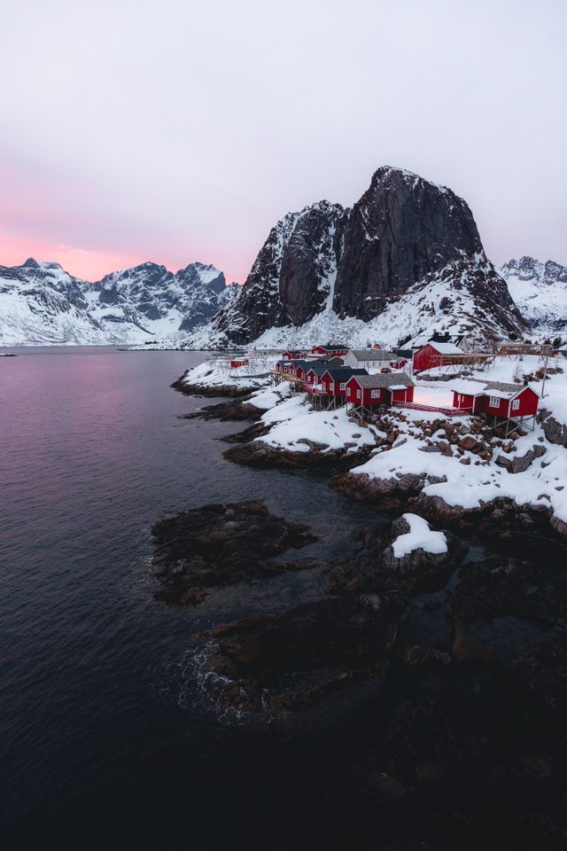 Case di legno rosse si ergono su una costa rocciosa e innevata accanto ad acque scure, con montagne innevate sotto un cielo rosa.