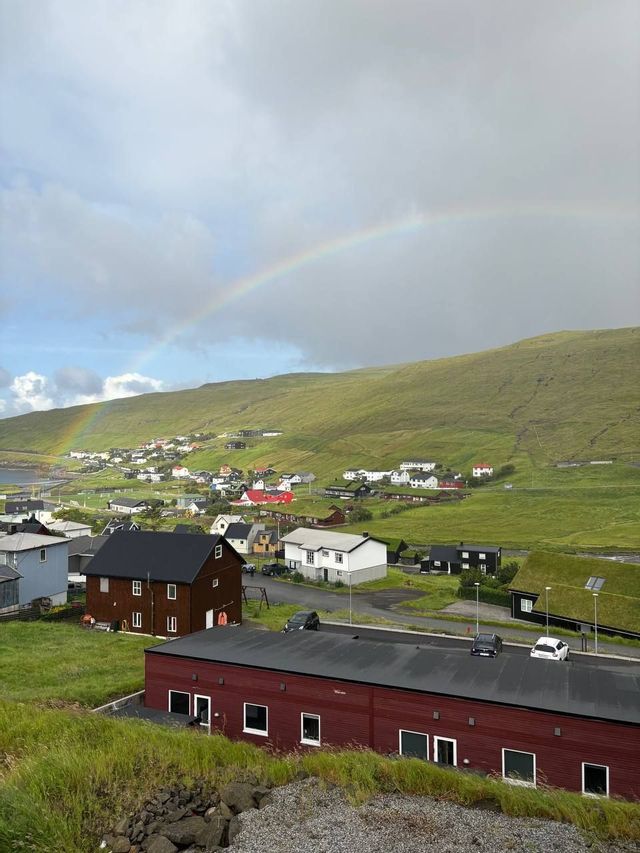Un arcobaleno si inarca su un piccolo villaggio adagiato ai piedi di una grande collina verde sotto un cielo nuvoloso.