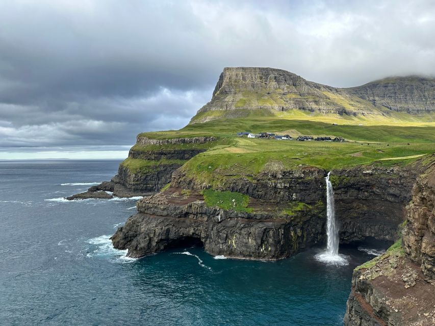 Una cascata si getta da una scogliera verde nell'oceano, con un piccolo villaggio visibile in cima alla scogliera sotto un cielo nuvoloso.