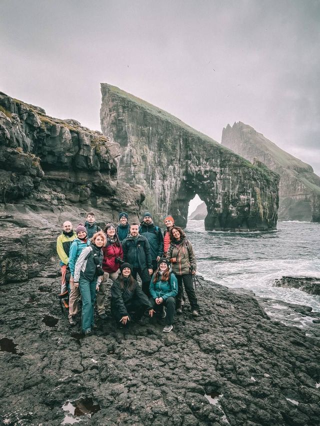 Un gruppo WeRoad in posa su una costa rocciosa, con grandi faraglioni e un arco naturale alle spalle, sotto un cielo nuvoloso.