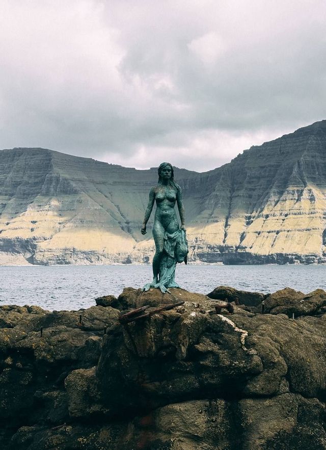 Una statua di bronzo di una selkie che tiene la sua pelle di foca si erge su rocce in riva al mare, di fronte a una scogliera montuosa stratificata.