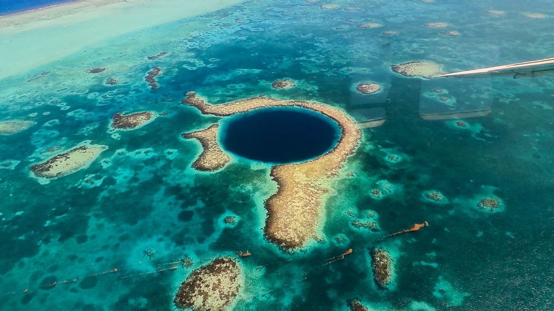 Veduta aerea di un grande buco blu circolare circondato da una barriera corallina in acque oceaniche turchesi, con l'ala di un aereo visibile.