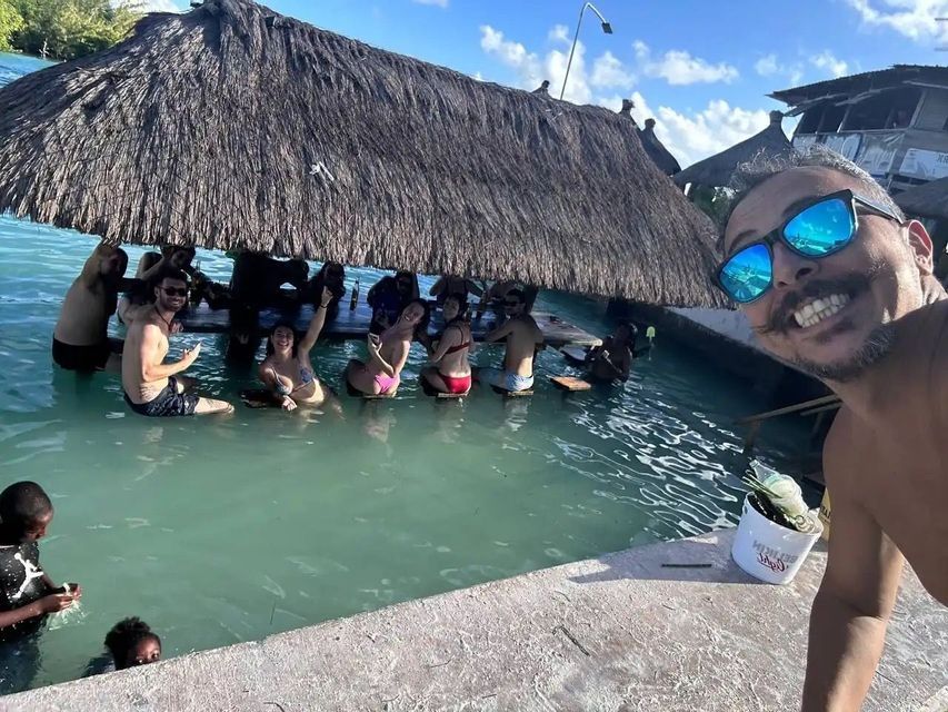 Un uomo scatta un selfie mentre un gruppo WeRoad si gode un momento di relax in un bar a bordo piscina con tetto di paglia.