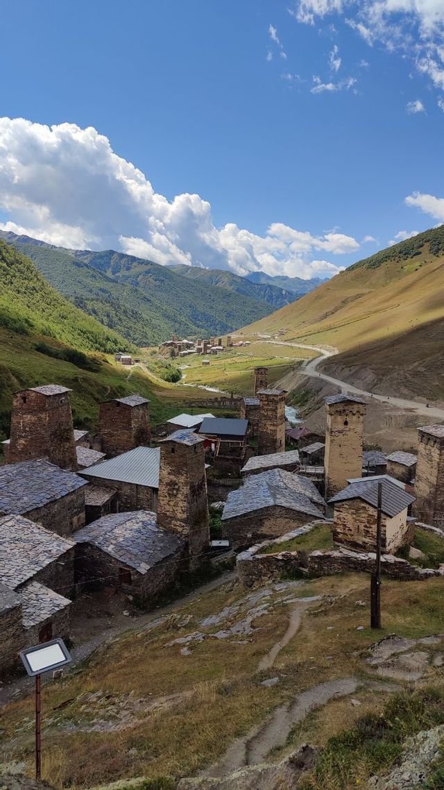 Un borgo di case in pietra e torri adagiato in una verde valle montana sotto un cielo parzialmente nuvoloso.
