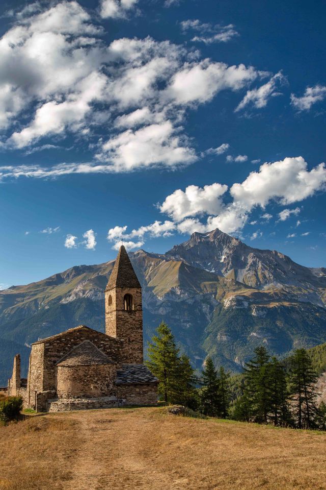 Una rustica chiesa in pietra con campanile si erge su una collina, con una vasta catena montuosa sullo sfondo, sotto un cielo azzurro e nuvoloso.