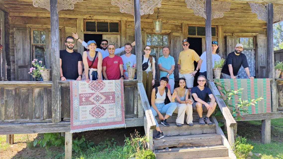 Un gruppo WeRoad in posa per una foto sul portico e sui gradini di una casa rustica in legno, con in mano un tappeto a motivi.