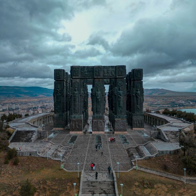 Veduta aerea di un imponente monumento con alte colonne scolpite, mentre persone camminano sulla sua piazza di pietra sotto un cielo nuvoloso.
