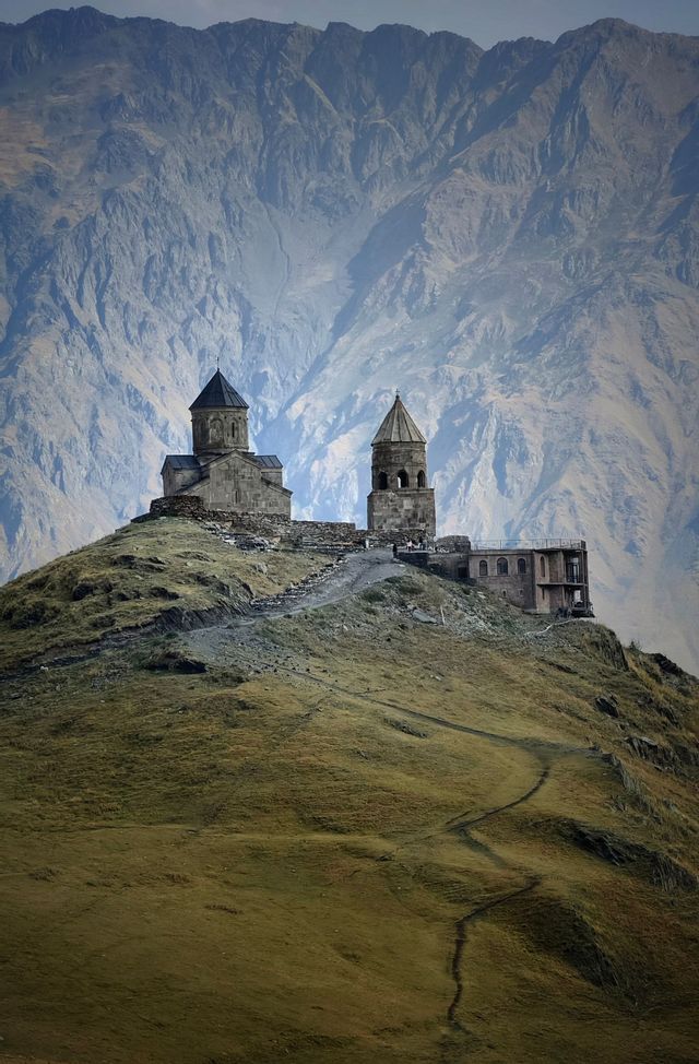 Una chiesa di pietra e un campanile si stagliano su una collina erbosa, con un'imponente e aspra catena montuosa a far da sfondo.