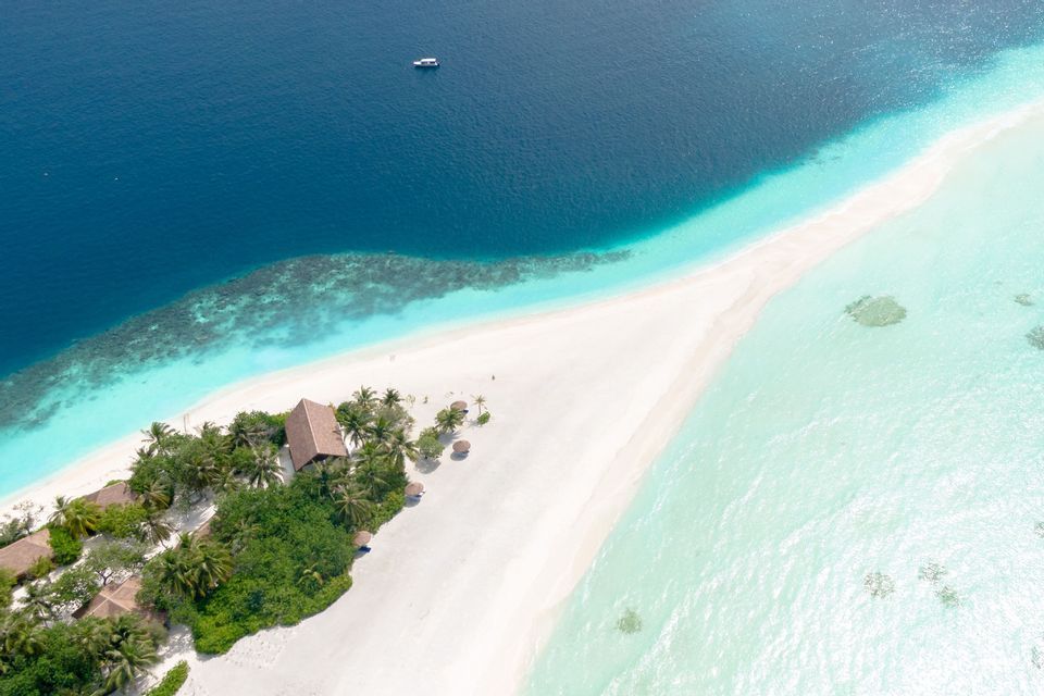 Veduta aerea di un lembo di sabbia bianca con palme ed edifici, che divide l'oceano blu intenso dalle acque turchesi poco profonde.