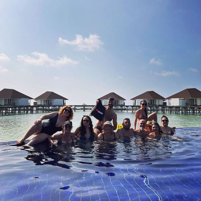 Un gruppo WeRoad in viaggio posa in una piscina a sfioro, con bungalow sull'acqua visibili sullo sfondo sotto un cielo azzurro.