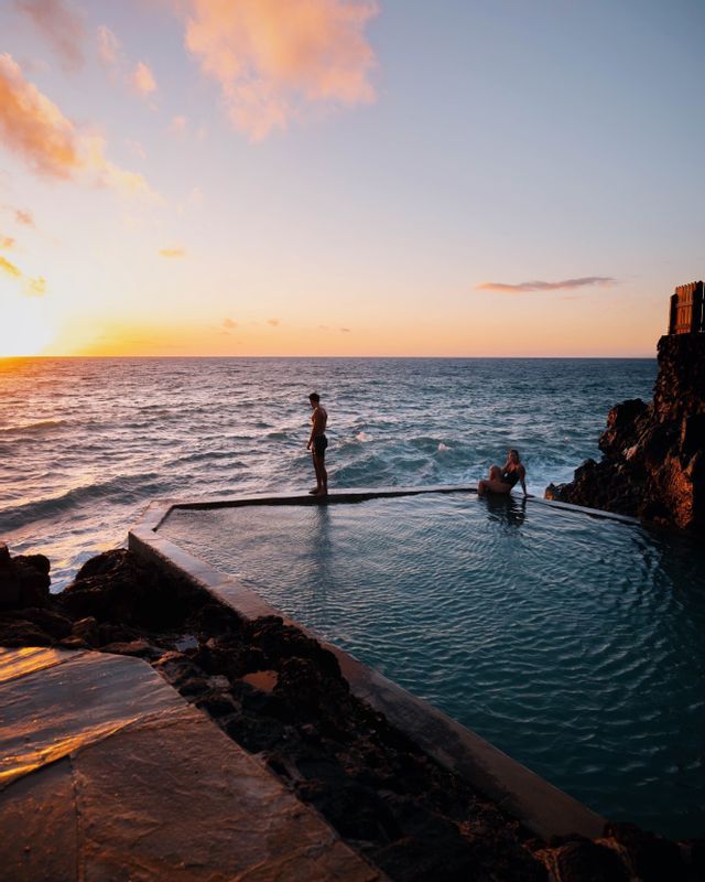 Un uomo in piedi e una donna seduta a bordo di una piscina a sfioro con vista sull'oceano, durante un tramonto colorato.