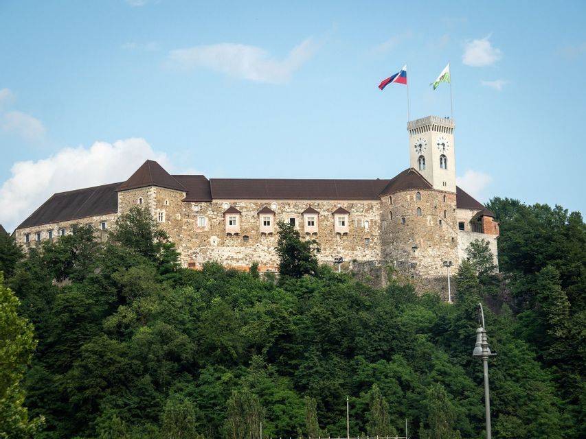 Un gran castillo de piedra con una destacada torre del reloj se alza sobre una exuberante colina arbolada bajo un cielo azul.