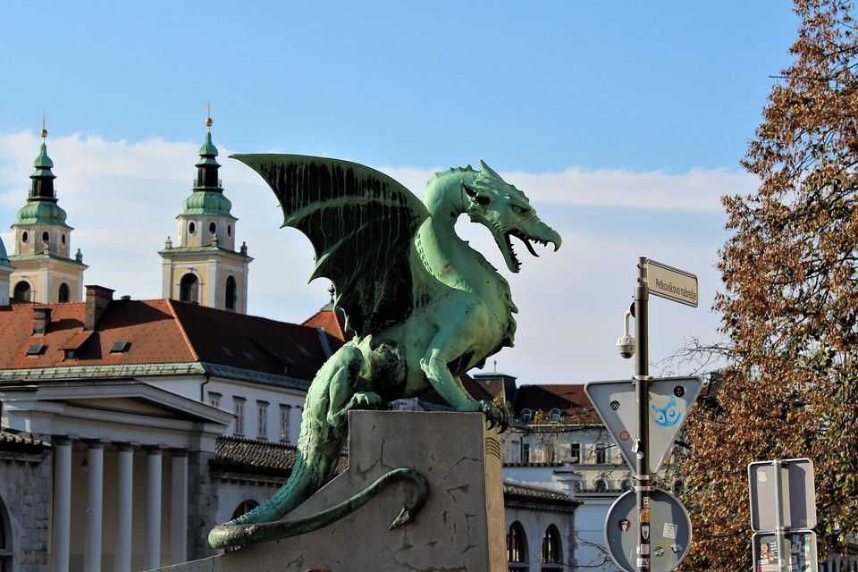 Una estatua de dragón verde con las alas extendidas se asienta sobre un pedestal, con edificios históricos y dos torres de iglesia al fondo.