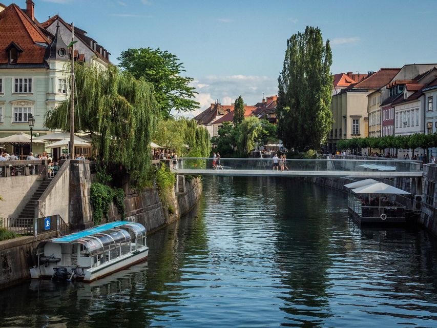 Un moderno puente peatonal de cristal cruza un canal de la ciudad, flanqueado por edificios históricos, cafés ribereños y barcos turísticos.