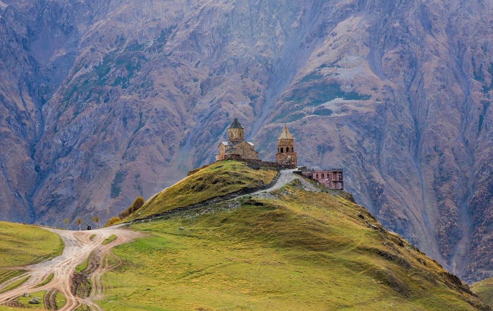Una storica chiesa in pietra e un campanile appollaiati su una collina erbosa di fronte a una vasta catena montuosa rocciosa.
