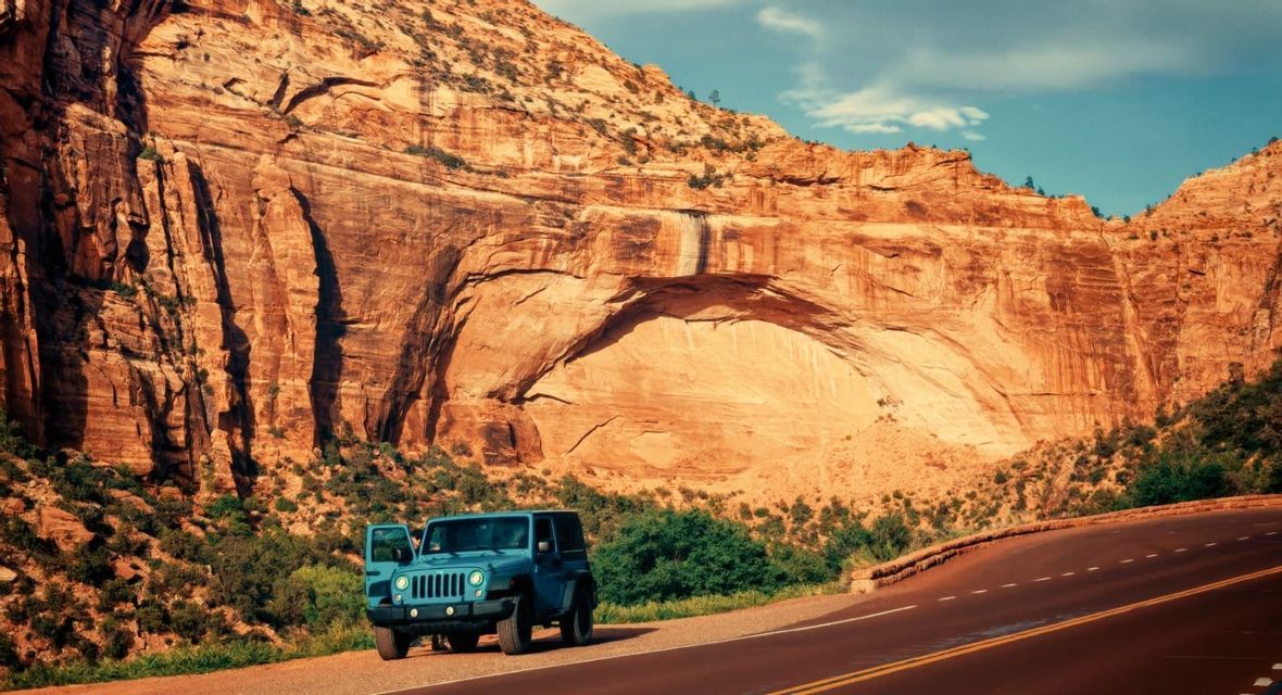 Una jeep blu con la portiera aperta è parcheggiata a bordo strada accanto a una grande formazione rocciosa di canyon rosso.
