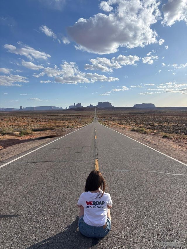 Un capogruppo WeRoad è seduto su una lunga strada dritta che conduce a formazioni rocciose in un paesaggio desertico sotto un cielo blu.