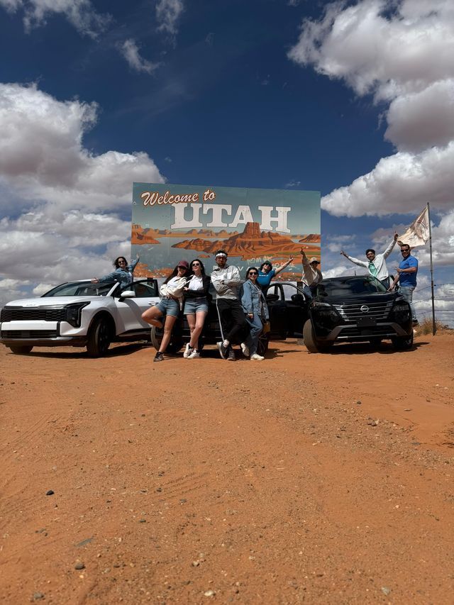 Un gruppo WeRoad posa per una foto con due auto davanti a un grande cartello 'Welcome to Utah' in un paesaggio desertico.