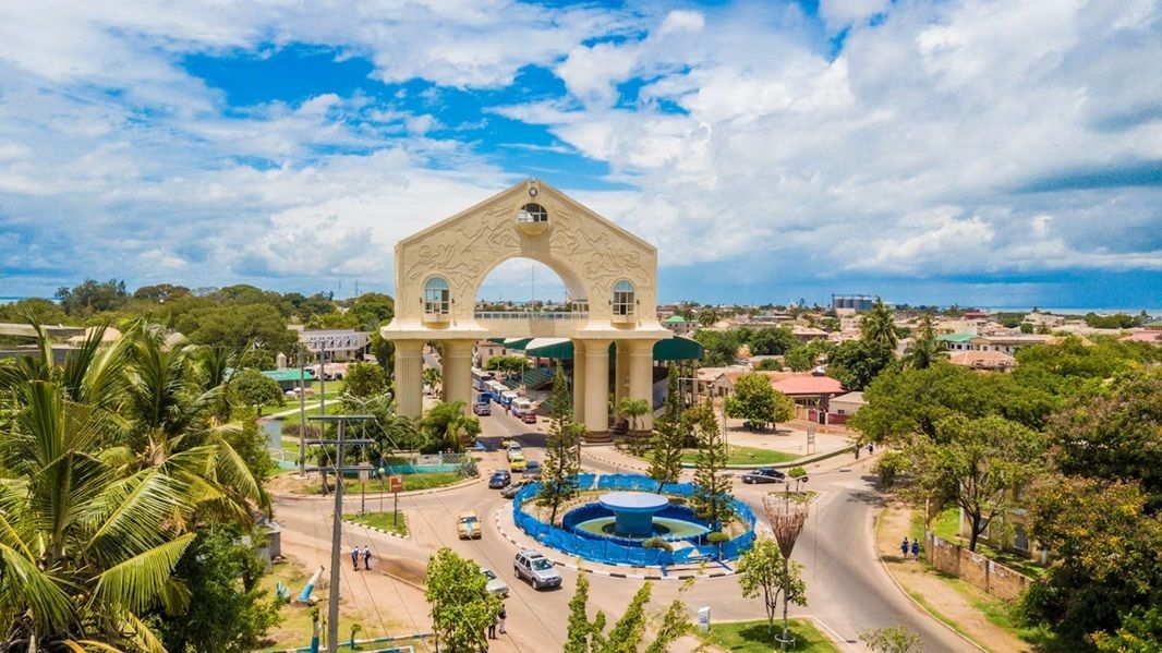 Una veduta aerea di un grande monumento ad arco sopra una rotonda stradale in una città costiera con alberi rigogliosi sotto un cielo azzurro nuvoloso.