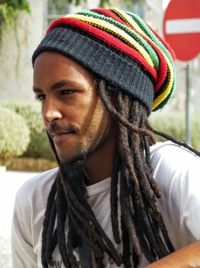 Un uomo con lunghi dreadlocks indossa un berretto a righe rosso, giallo e verde, mentre guarda di lato all'aperto.