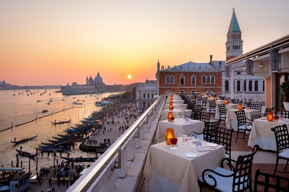 Set tables on a rooftop restaurant overlook a historic city's waterfront with boats and gondolas at sunset.