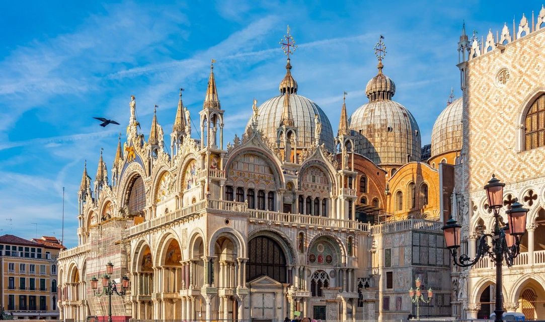 The detailed facade of an ornate basilica with multiple domes and spires stands against a bright blue sky.