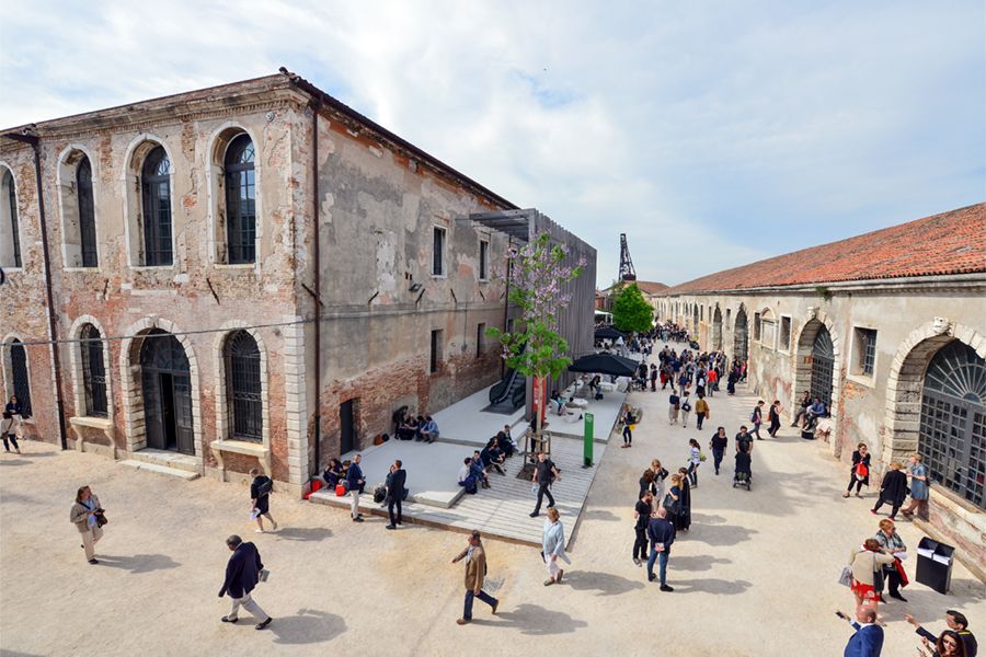 A high-angle shot of people walking and relaxing in a large courtyard between two old, weathered brick buildings under a cloudy sky.