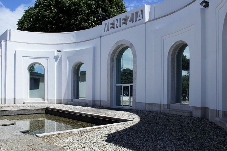A modern white building with a curved facade, arched windows, and the word 'VENEZIA' carved above, next to a gravel courtyard.