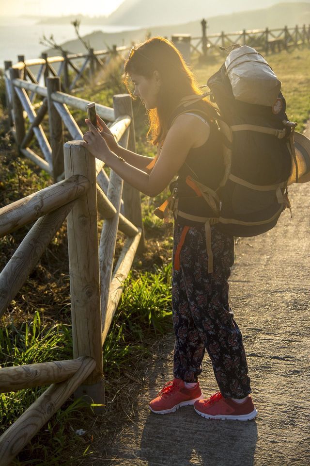 Un'escursionista con un grande zaino si ferma vicino a una staccionata di legno per scattare una foto del tramonto con il suo telefono.