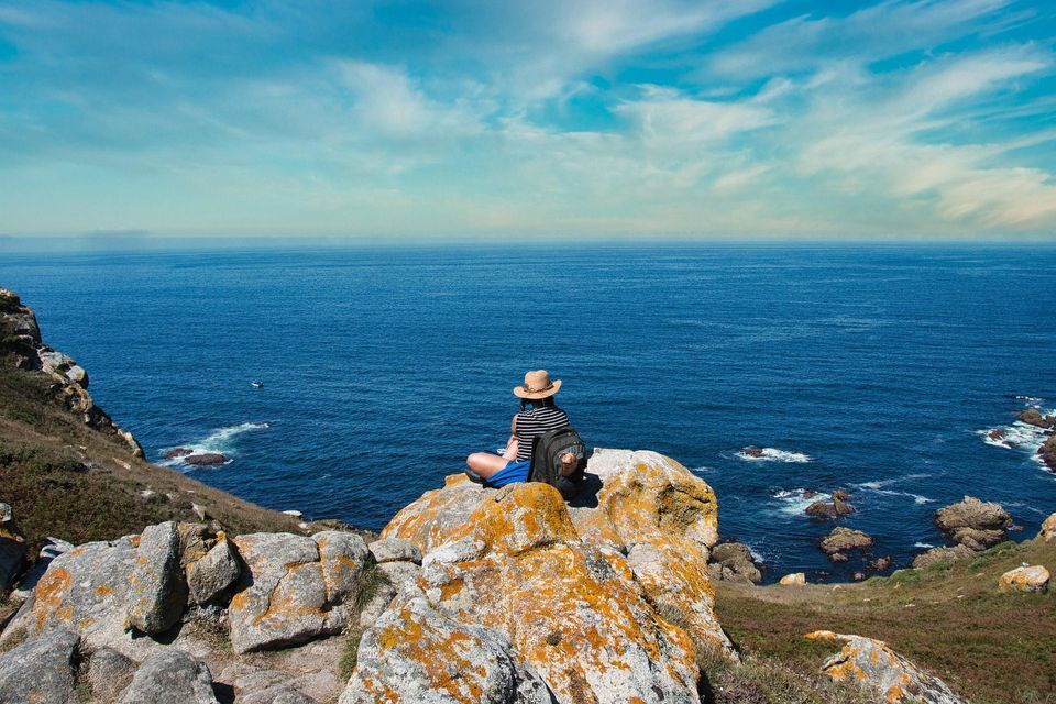 Una persona con un cappello di paglia e uno zaino siede su una scogliera rocciosa affacciata sull'oceano blu.